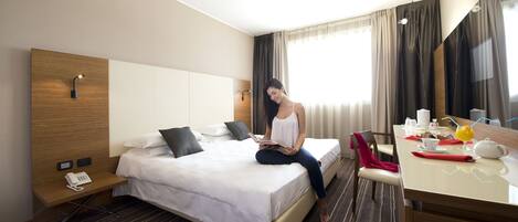 A hotel room with a bed, bedside table, a desk with a chair, a telephone, and a window with curtains.