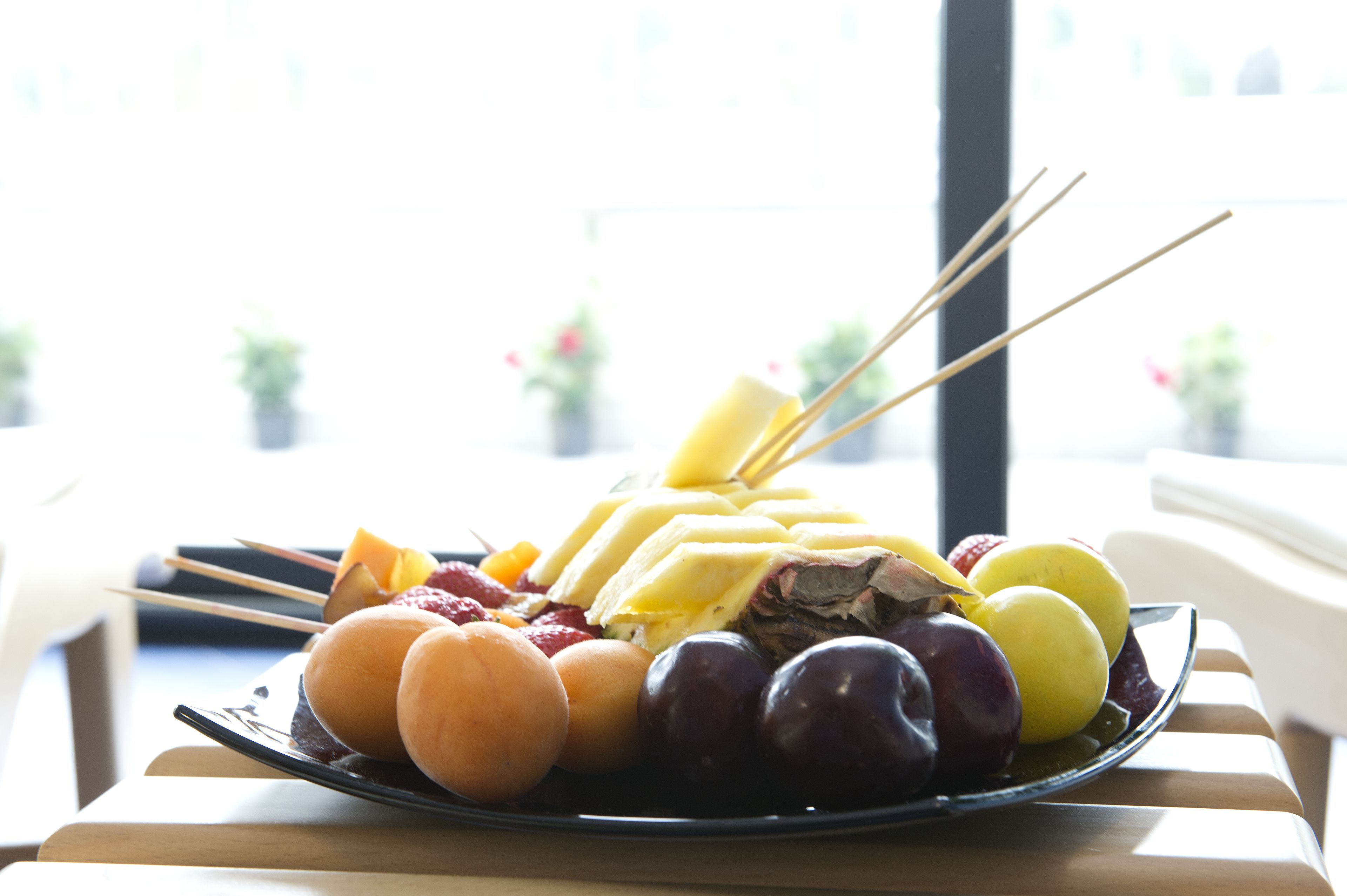 Une assiette remplie de fruits variés et une banane sont posées sur une table, avec vue sur un bâtiment à l'extérieur.