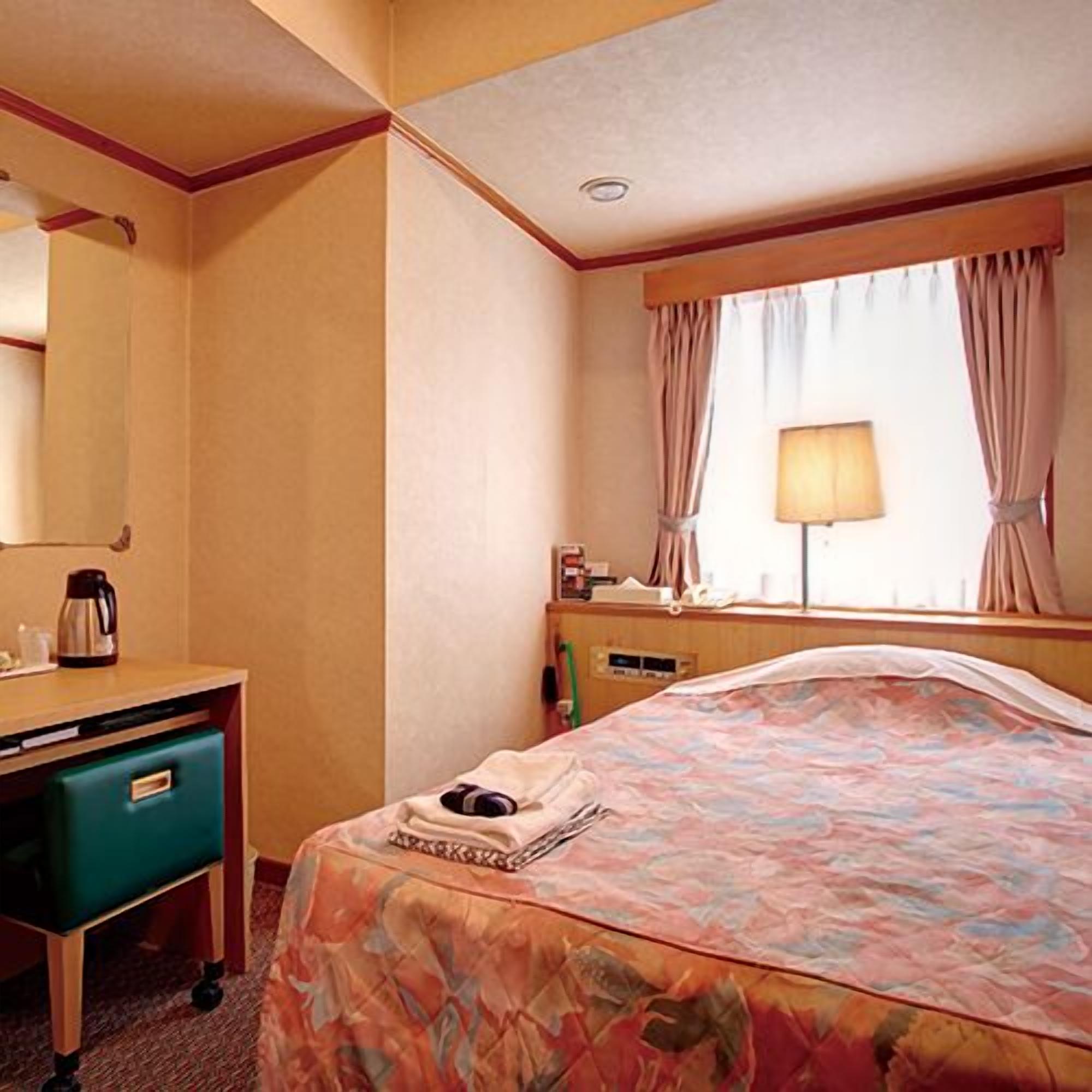 A hotel room with a bed, a desk with a coffee maker, a mirror, a window with curtains, and a lamp.