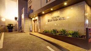 The exterior of Okinawa Nishi Plaza Hotel with a prominent entrance, a small garden area with plants, and a security booth. - Okinawa Sunplaza Hotel (Naha)