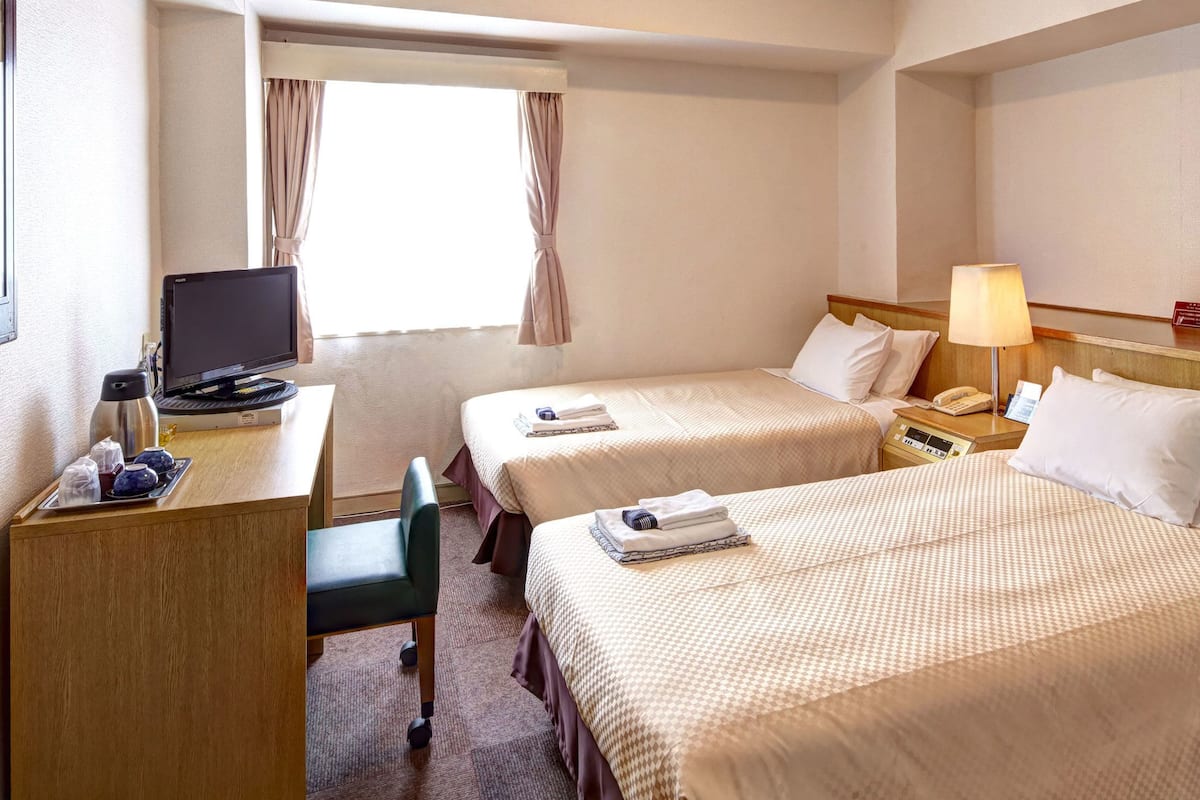 A hotel room with two beds, a desk with a computer, a chair, and a bedside table with a lamp.