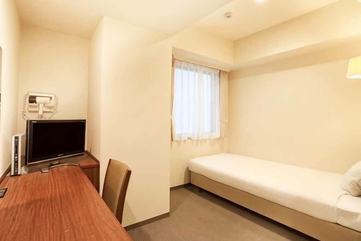 A small hotel room with a wooden desk, a television, a single bed, and a window with curtains.