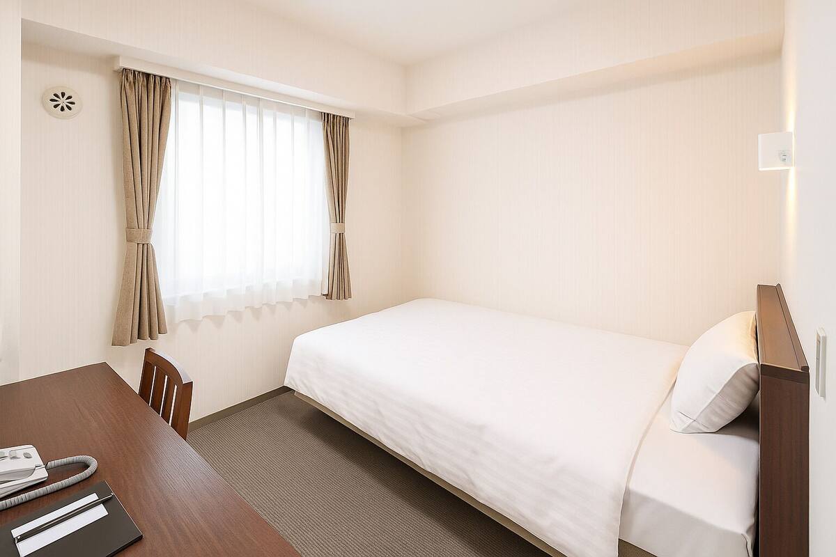 A hotel room with a single bed, a wooden desk, and a window with curtains.