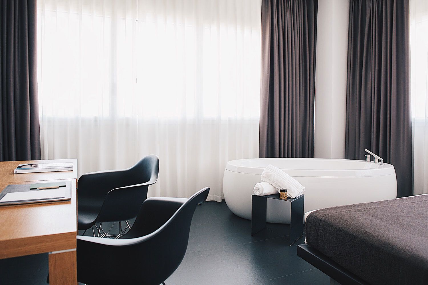 Una habitación de hotel moderna con escritorio, dos sillas, un sofá, una bañera y un ventanal con cortinas.