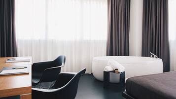 Una habitación de hotel moderna con escritorio, dos sillas, un sofá, una bañera y un ventanal con cortinas.