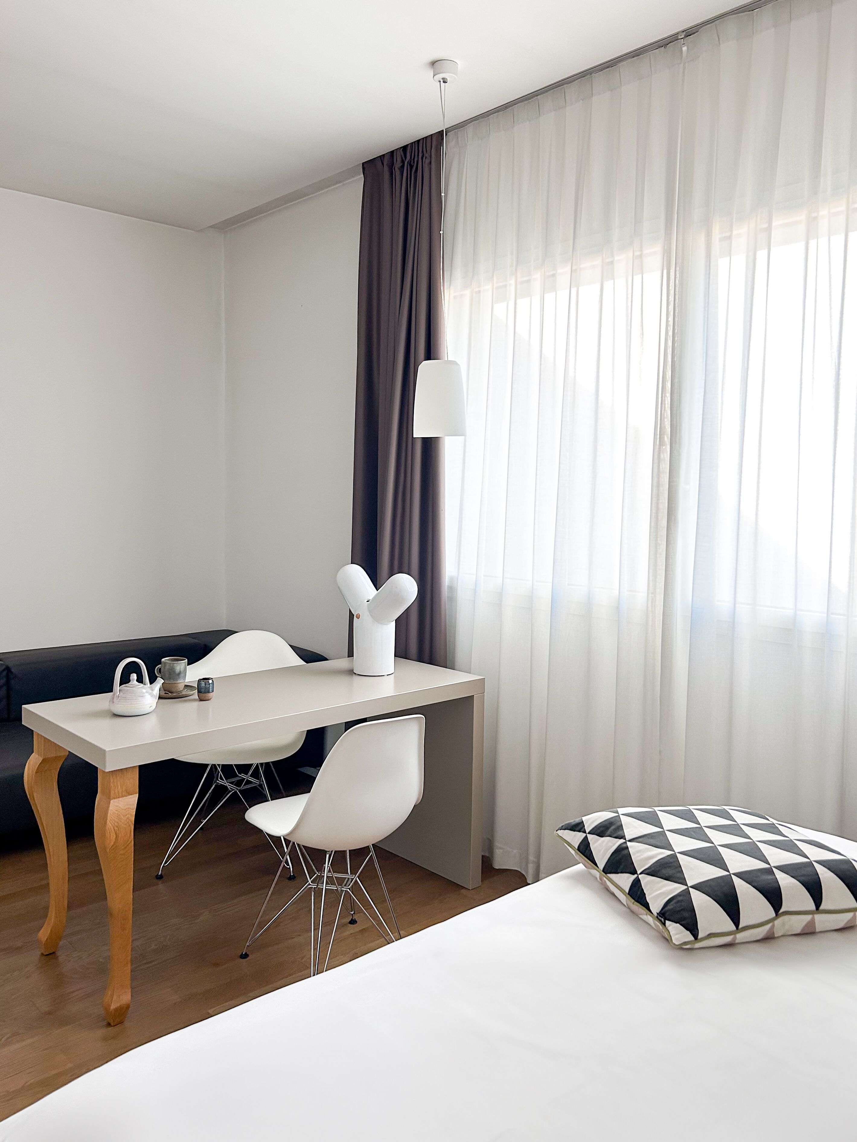 A modern hotel room with a bed, a desk with chairs, a sofa, and a large window with curtains.