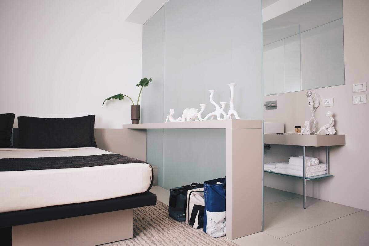 A modern bedroom with a bed, a bedside plant, a mirror, and a shelf with towels and toiletries.