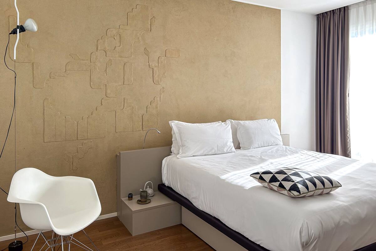 A modern hotel room with a large bed, white bedding, a geometric wall design, a white chair, and a bedside table with a kettle.