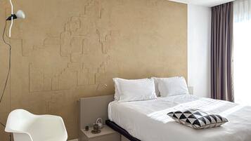 Habitación de hotel moderna con una cama grande, ropa de cama blanca, diseño geométrico en la pared, una silla blanca y una mesita de noche con hervidor.