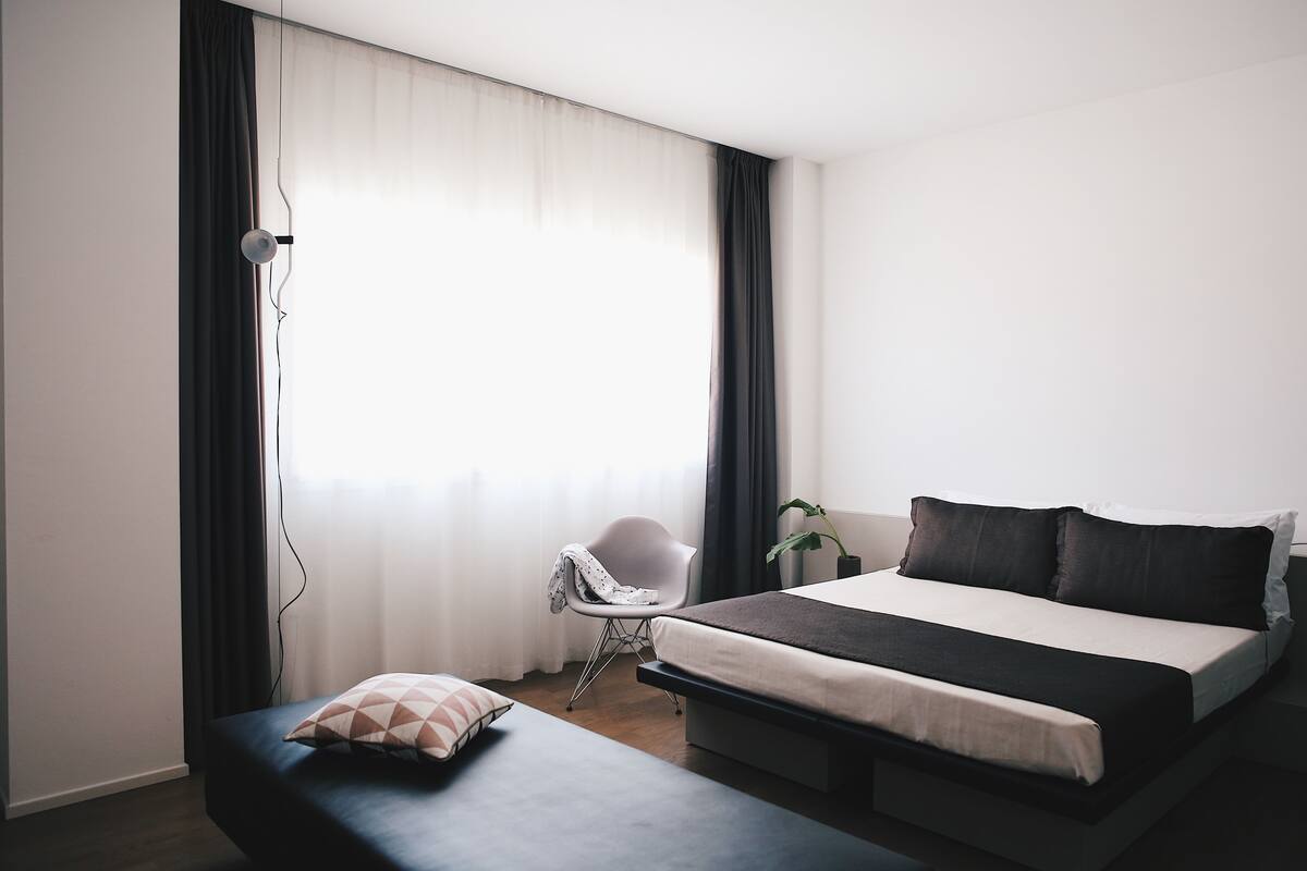 A modern hotel room with a bed, bedside table, chair, and a large window with curtains.
