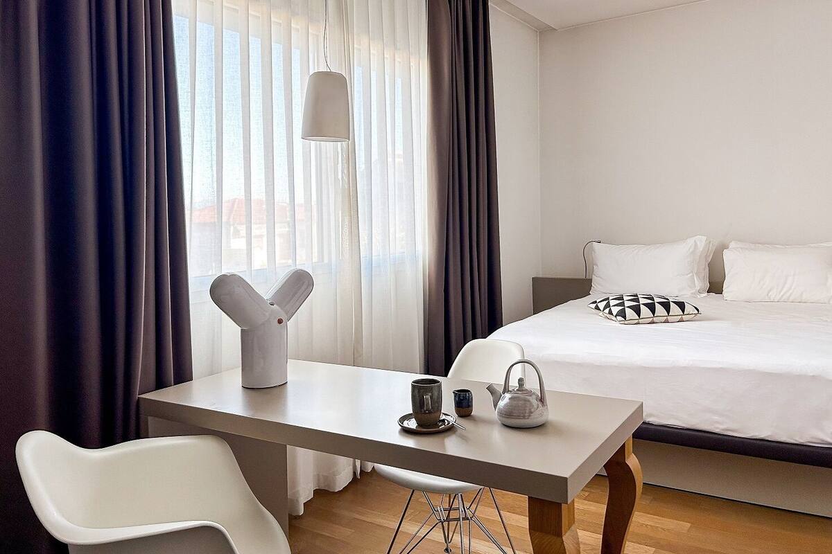 A hotel room with a bed, a desk with a chair, a kettle, and a teapot.