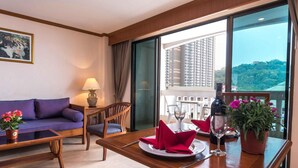 Habitación de hotel con balcón, una mesa de comedor con mantel individual rojo, una copa de vino y una botella. Hay un sofá y una silla en la habitación, y una planta en maceta sobre la mesa.