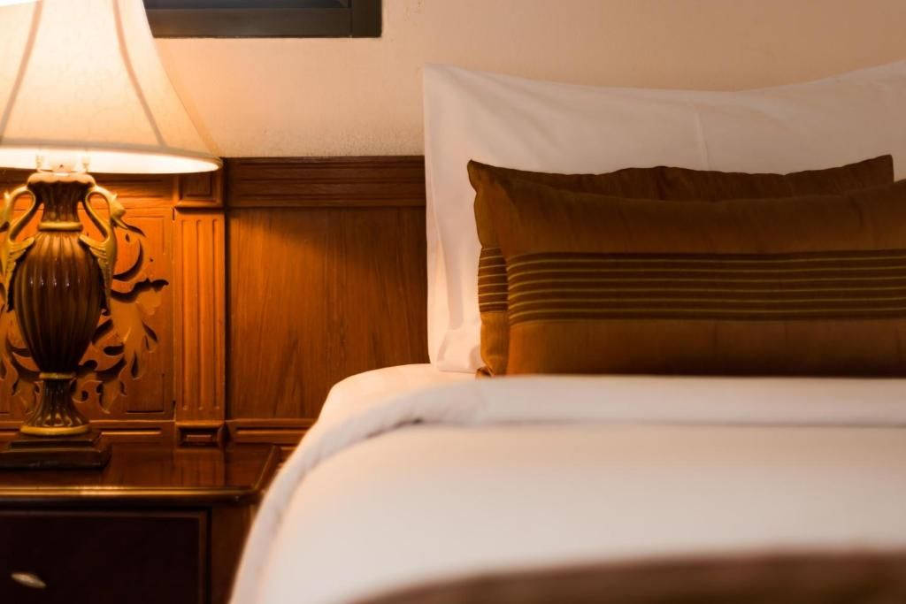 Een net opgemaakt bed met een wit kussen en een bruin kussen, een houten nachtkastje met een lamp, en een muur met een houten paneelstructuur.