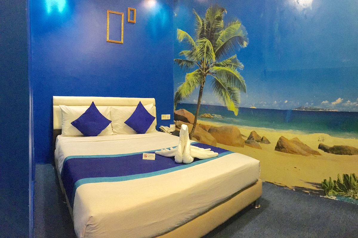 A hotel room with a bed, blue walls, a beach mural, and a palm tree.