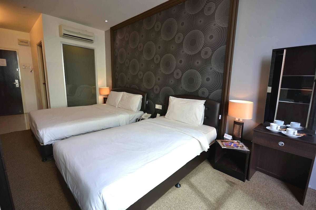 A hotel room with two beds, a dark patterned headboard, a desk with a mirror, and a small table with cups and saucers.