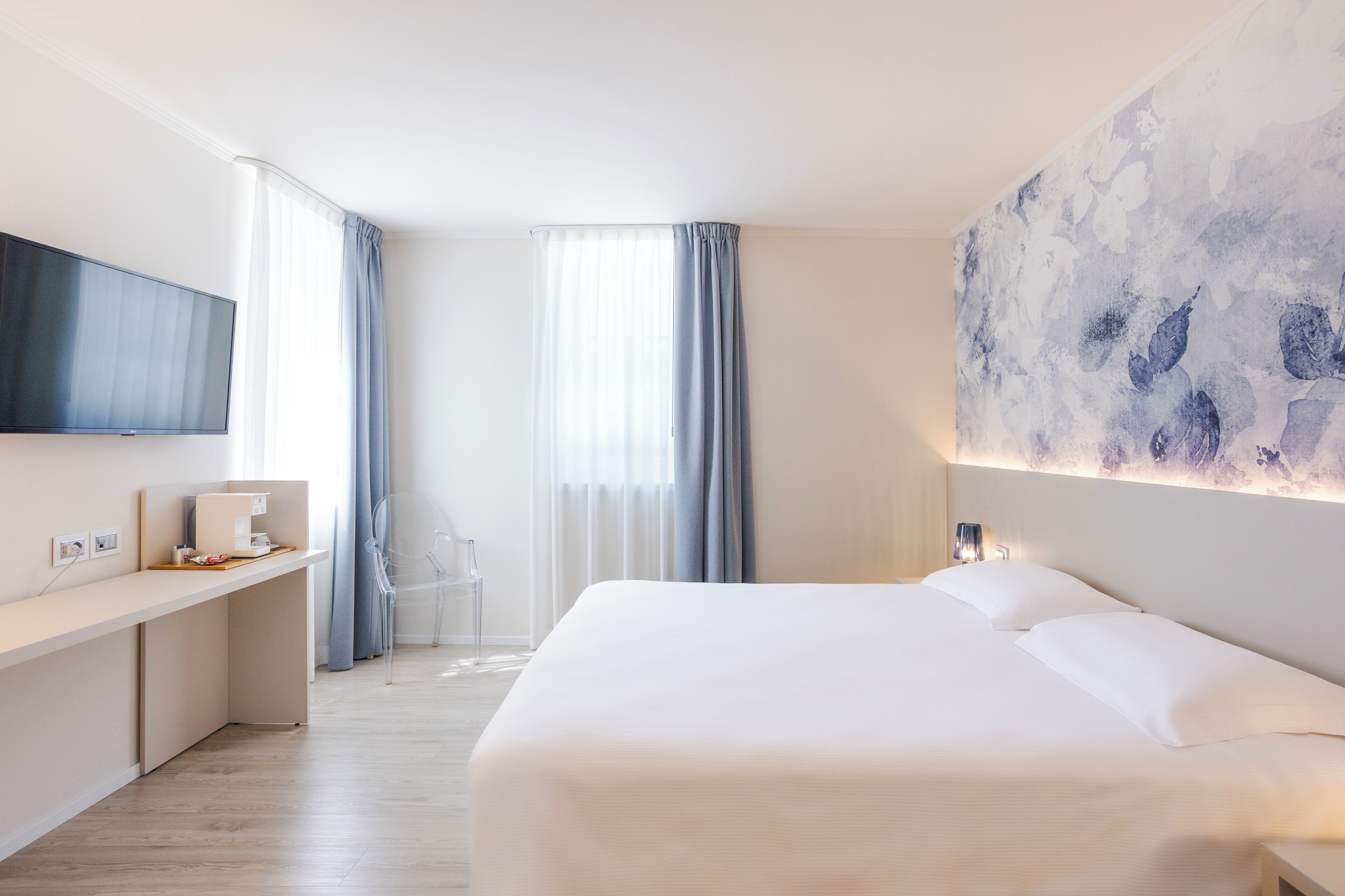 Una habitación de hotel moderna con una cama grande, un televisor de pantalla plana montado en la pared, un escritorio con una cafetera y un cuadro abstracto de gran tamaño en la pared.