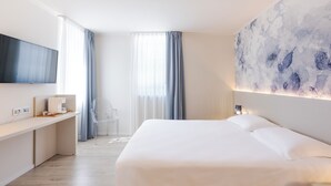 A modern hotel room with a large bed, a flat-screen TV mounted on the wall, a desk with a coffee machine, and a large abstract painting on the wall.