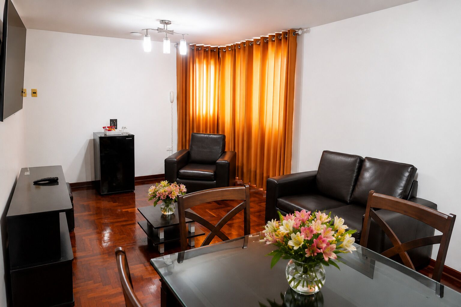 A modern living room with a glass-top dining table, two black leather sofas, a black coffee table, a black cabinet, and a vase with flowers.
