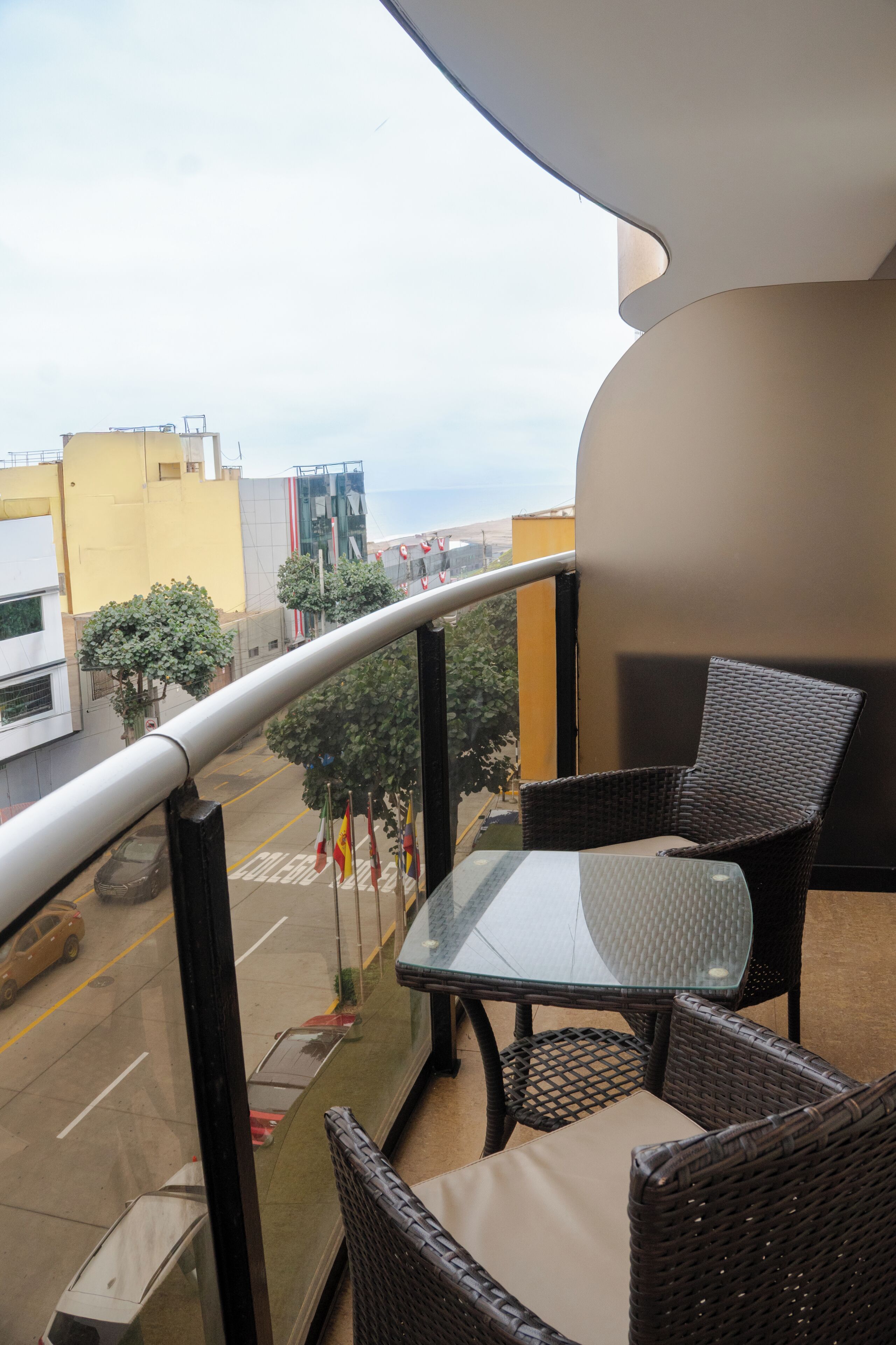 A balcony with wicker furniture, a glass table, and a view of the street and buildings.