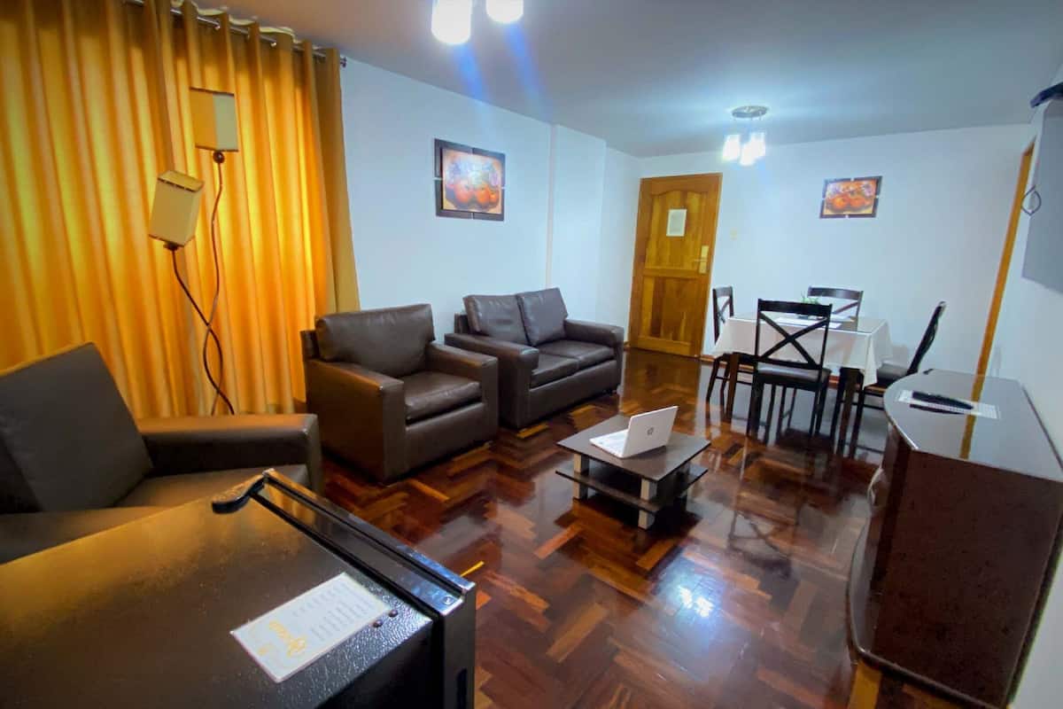 Una sala de estar amoblada con sofá, sillón, mesa de comedor y sillas. Hay una laptop sobre una mesita y un parlante a la derecha.
