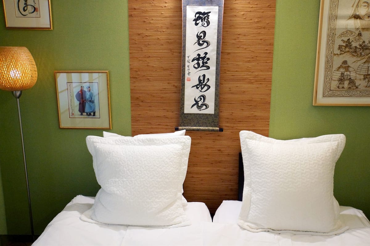 A hotel room with a wooden headboard, two white pillows, a framed calligraphy piece, and a traditional lamp.
