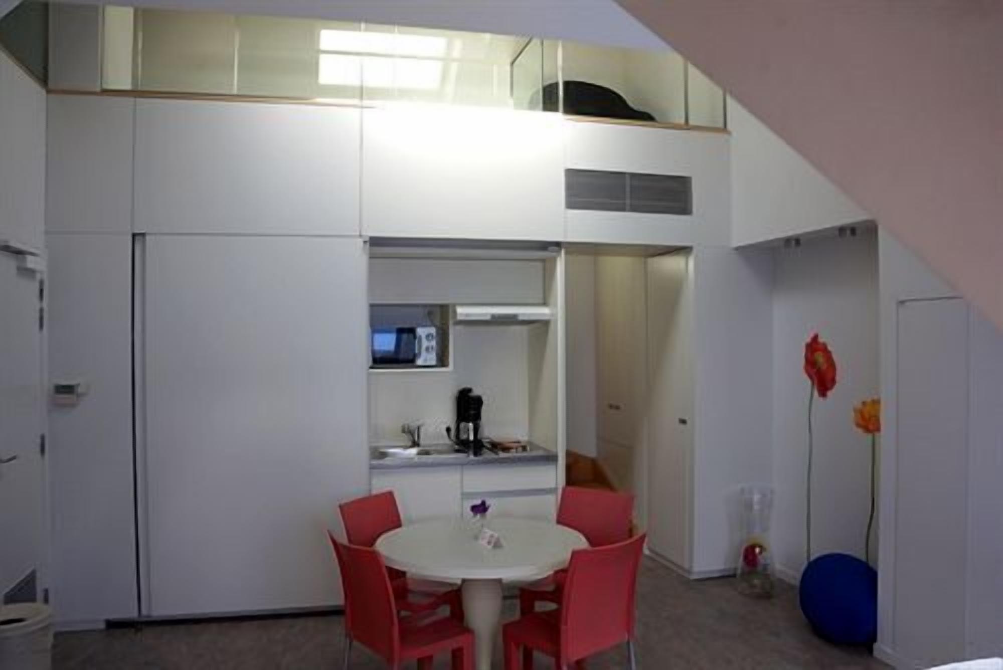 A compact room with a kitchenette, a dining area with red chairs, and a small window.