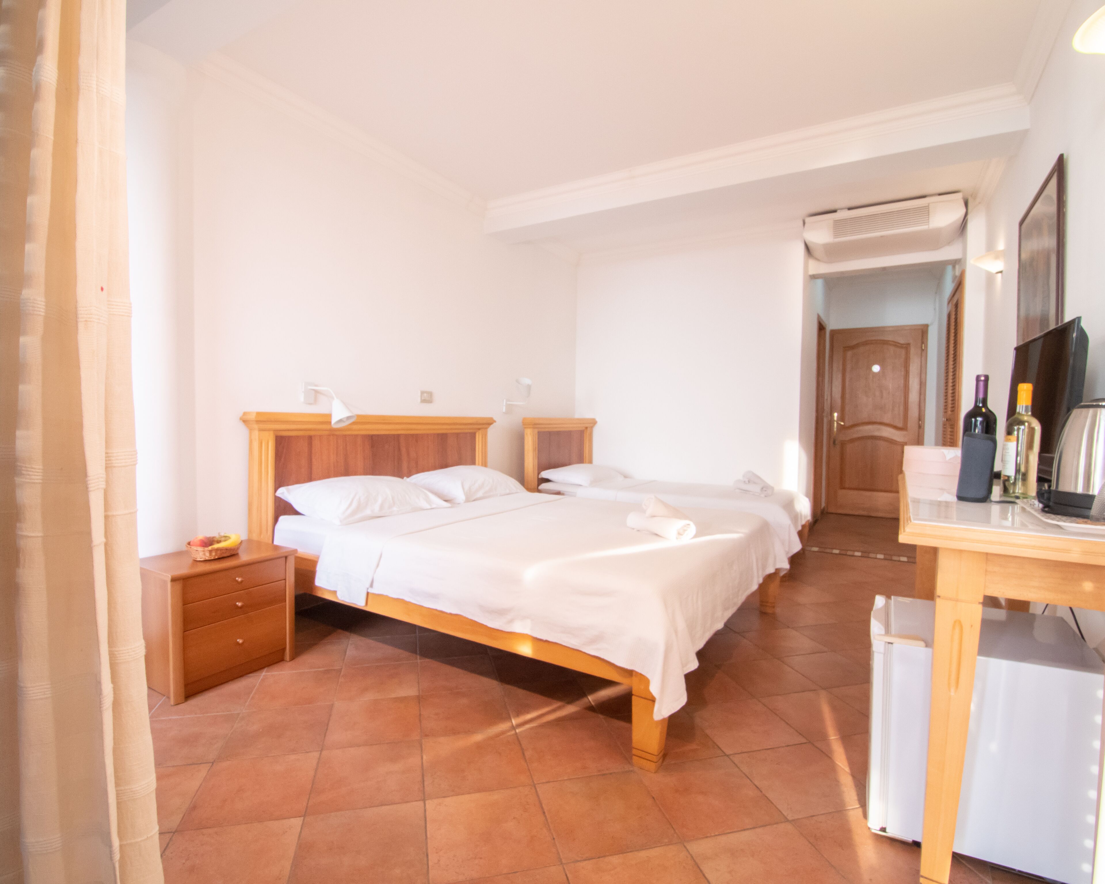 Un dormitorio con dos camas individuales, una mesita de noche de madera, un escritorio con una silla, un espejo y una puerta que da a otra habitación.