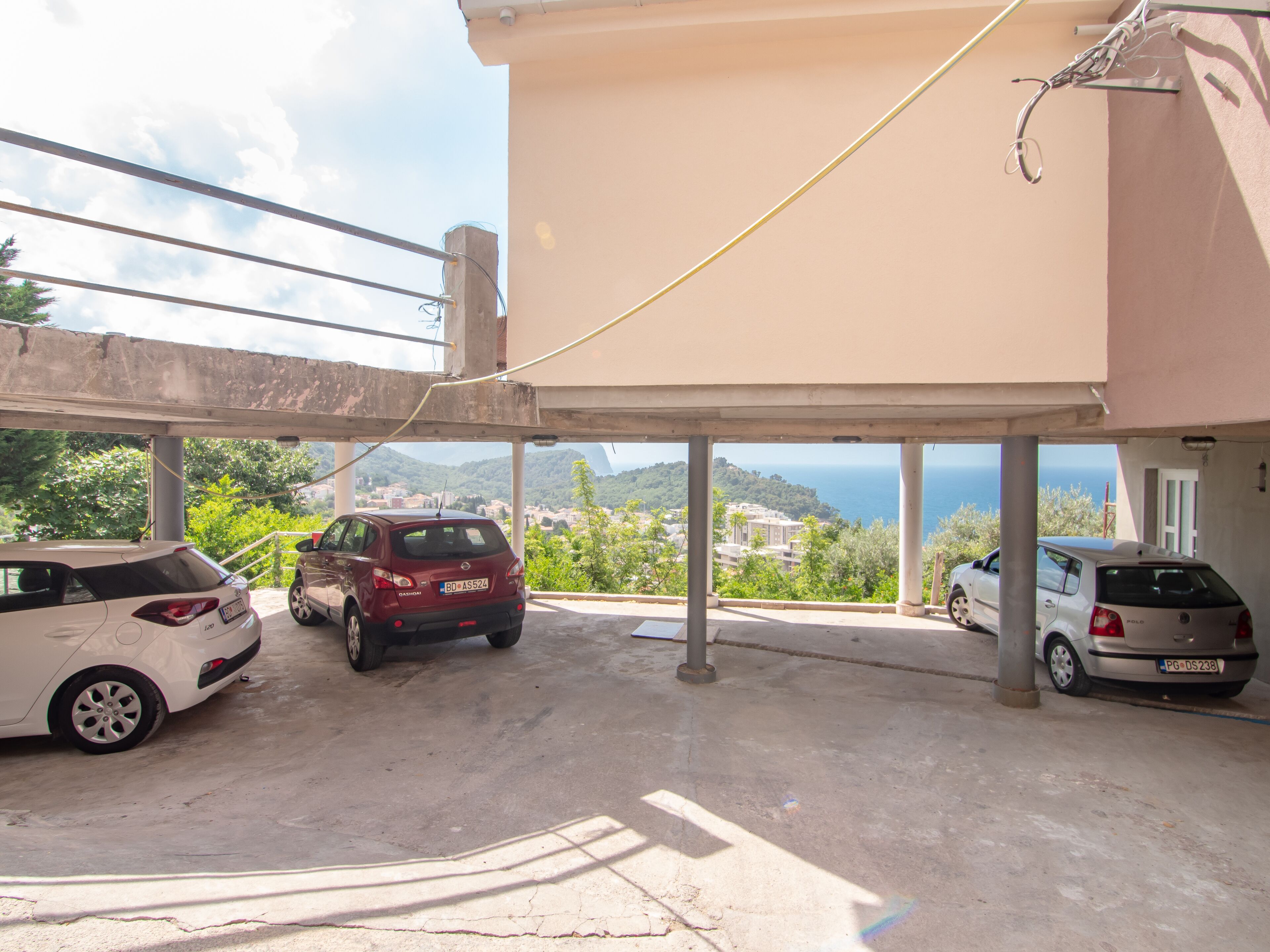 A covered parking area with three cars parked. The area is adjacent to a building with a view of a coastal landscape.