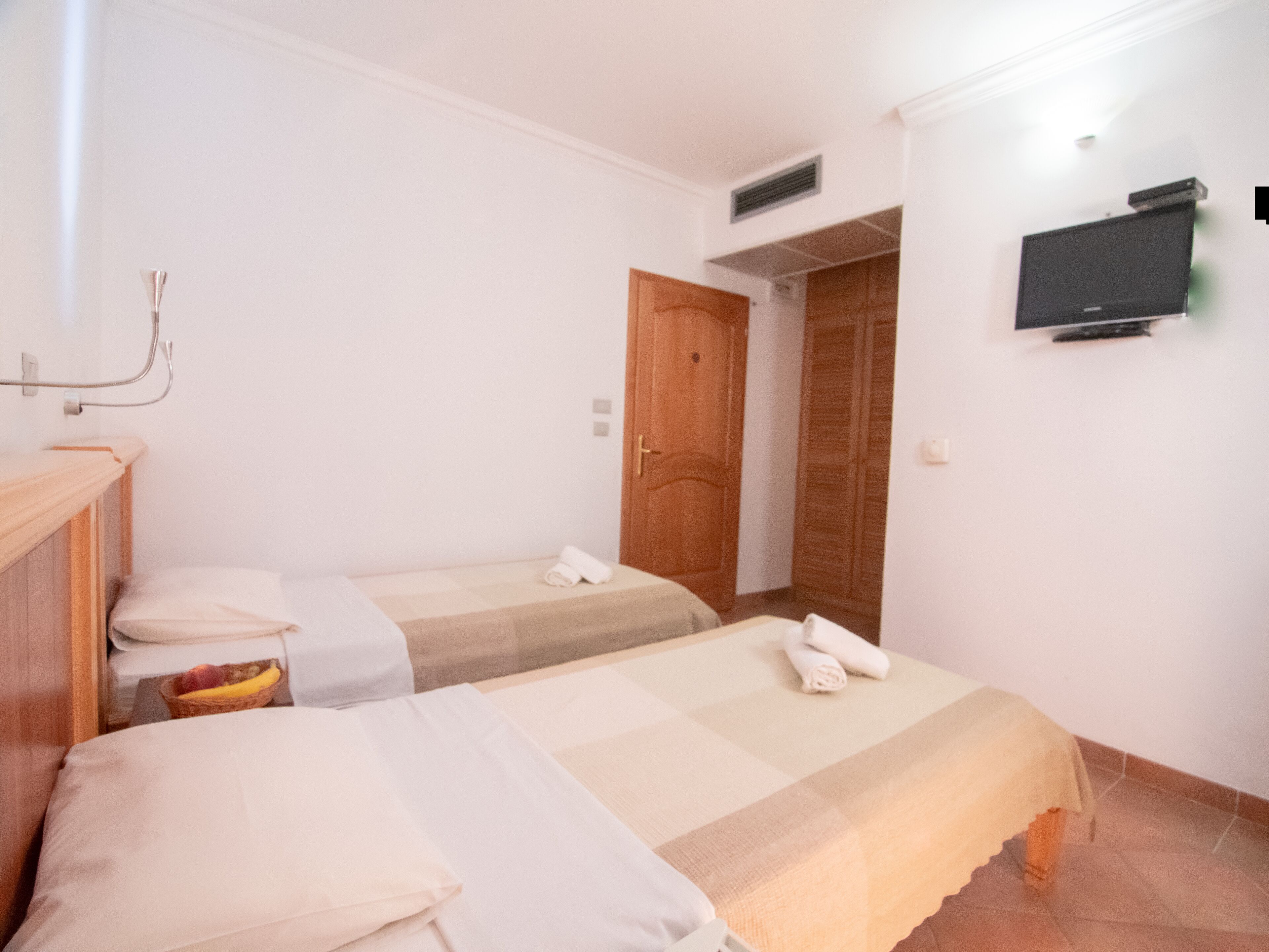 A hotel room with two single beds, a wooden wardrobe, a television mounted on the wall, and a door leading to another room.