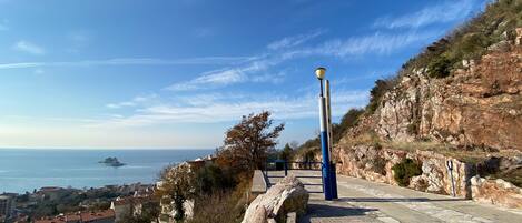 A paved pathway with a lamp post, rocky terrain, and a view of the sea and buildings in the distance.