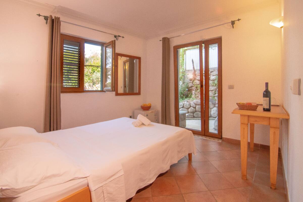 Ein Schlafzimmer mit einem Bett, einem Nachttisch, einem Tisch mit einer Flasche und Blick durch eine Glastür.