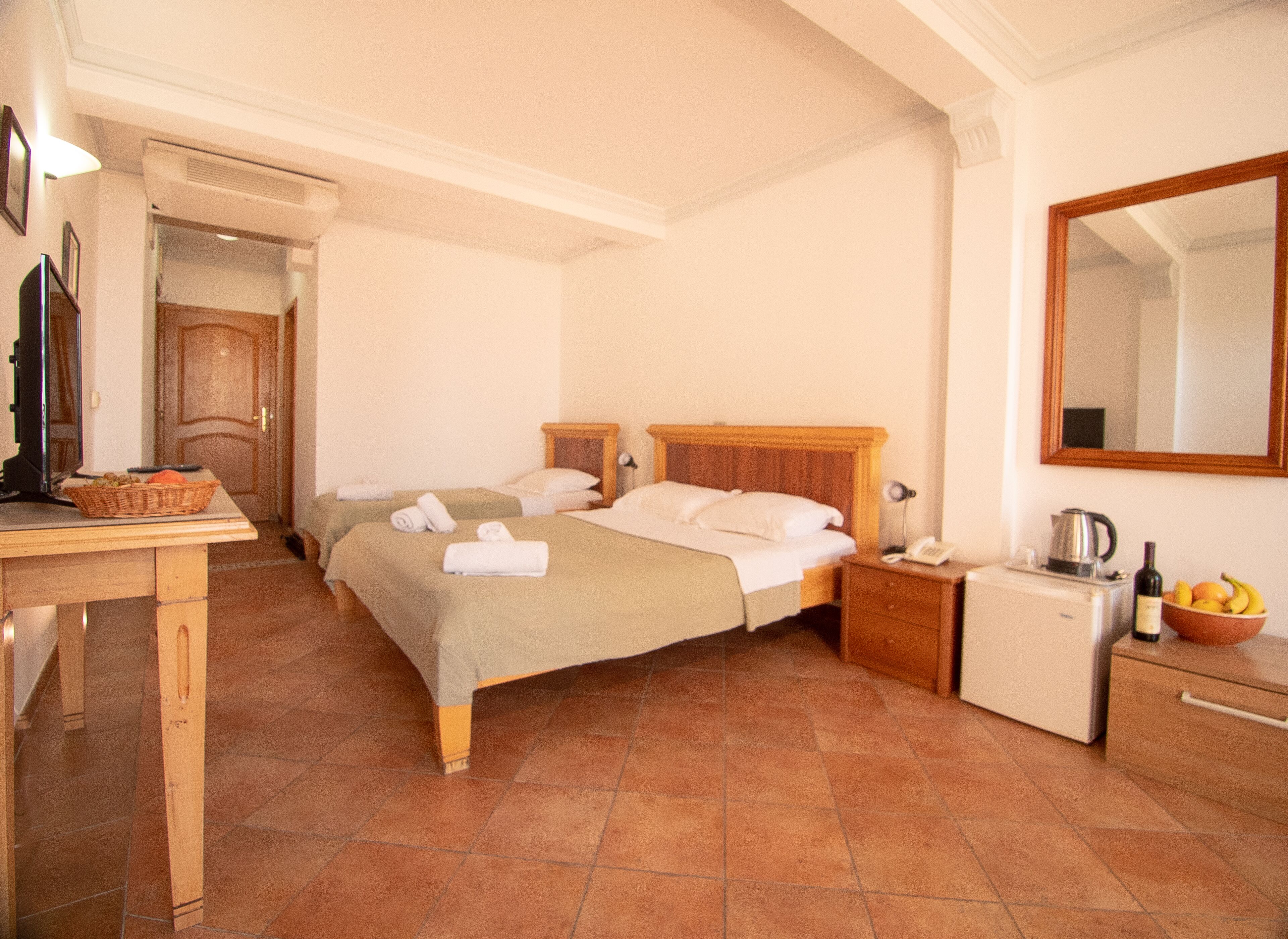 A hotel room with a bed, bedside tables, a desk with a basket, a small refrigerator, and a mirror on the wall.