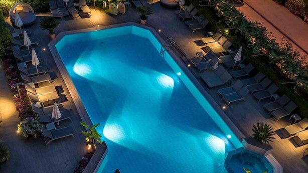 An aerial view of a rectangular swimming pool surrounded by lounge chairs and tables, with a wooden deck and potted plants. - Hotel Florida (Levico Terme)