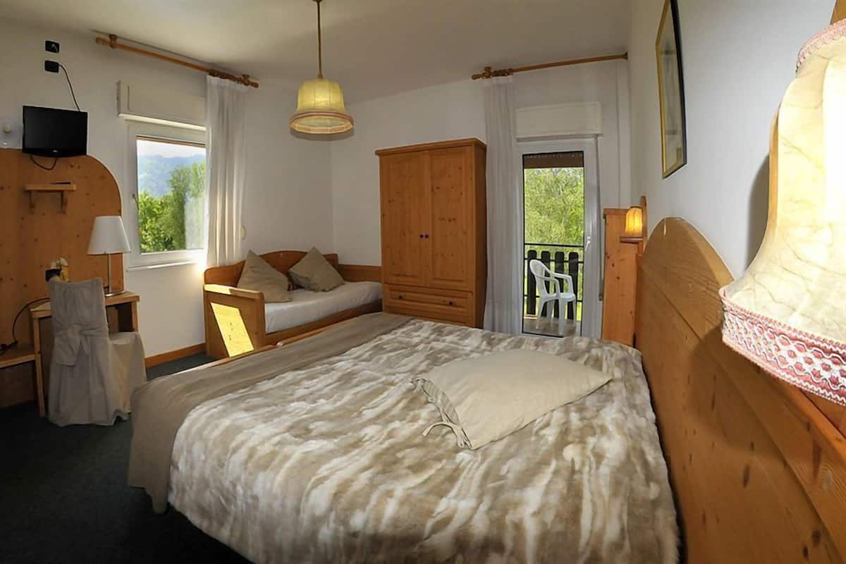 A bedroom with a bed, a wooden wardrobe, a desk, a chair, a TV, and a balcony with a chair.