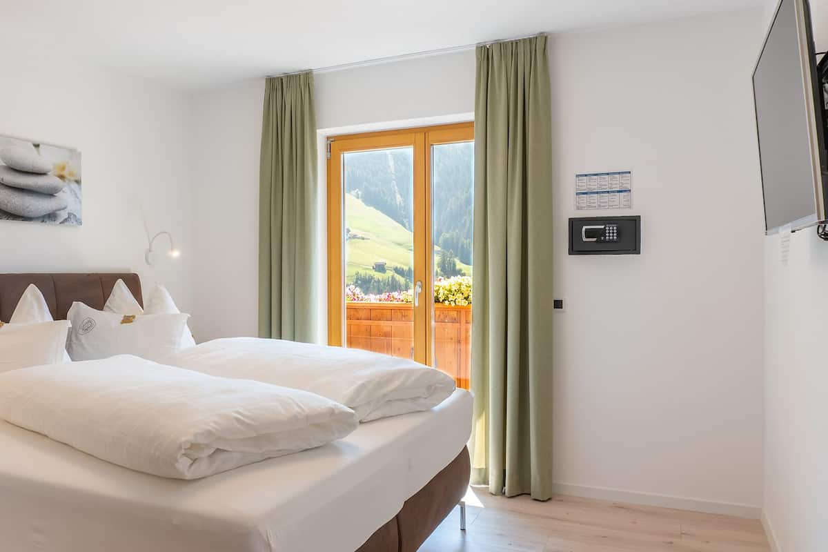 A bedroom with a bed, pillows, a painting, a window with curtains, and a wall-mounted control panel.