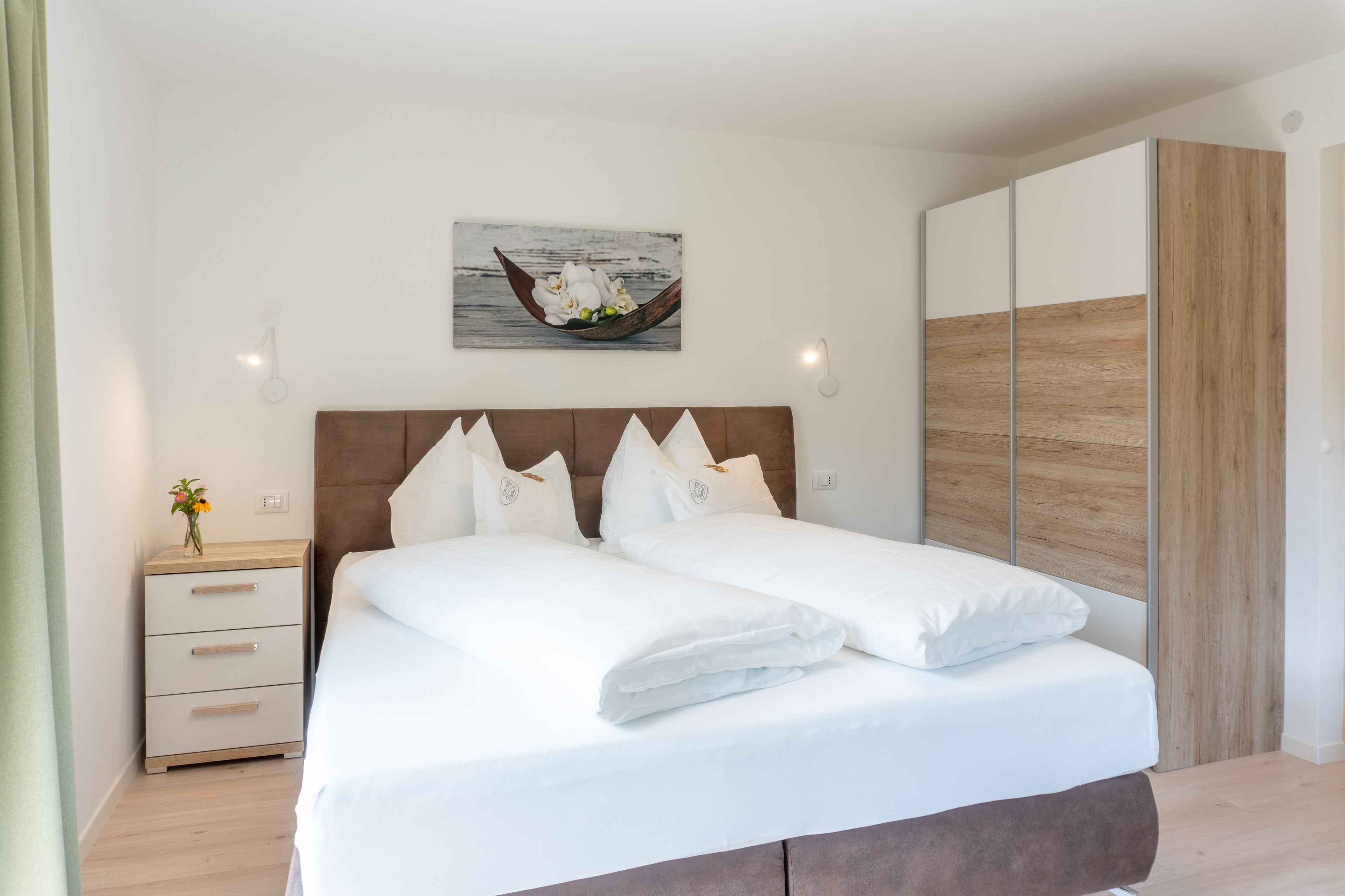 A neatly made bed with white linens and pillows, a wooden headboard, a bedside table with a vase of flowers, and a wall-mounted picture of a boat.