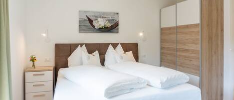 A neatly made bed with white linens and pillows, a wooden headboard, a bedside table with a vase of flowers, and a wall-mounted picture of a boat.