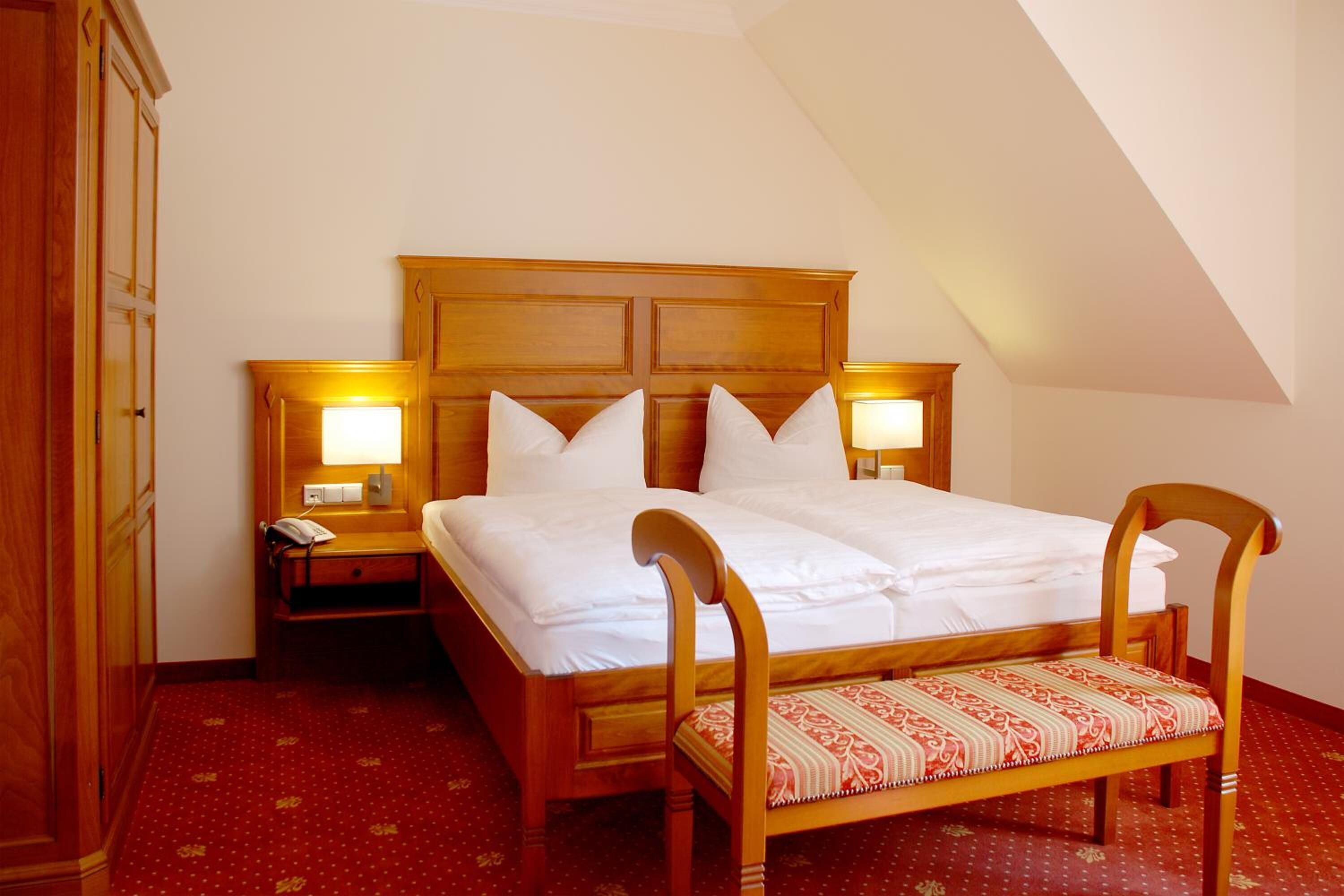 A hotel room with a wooden bed, two bedside tables with lamps, a wooden bench, and a red patterned carpet.