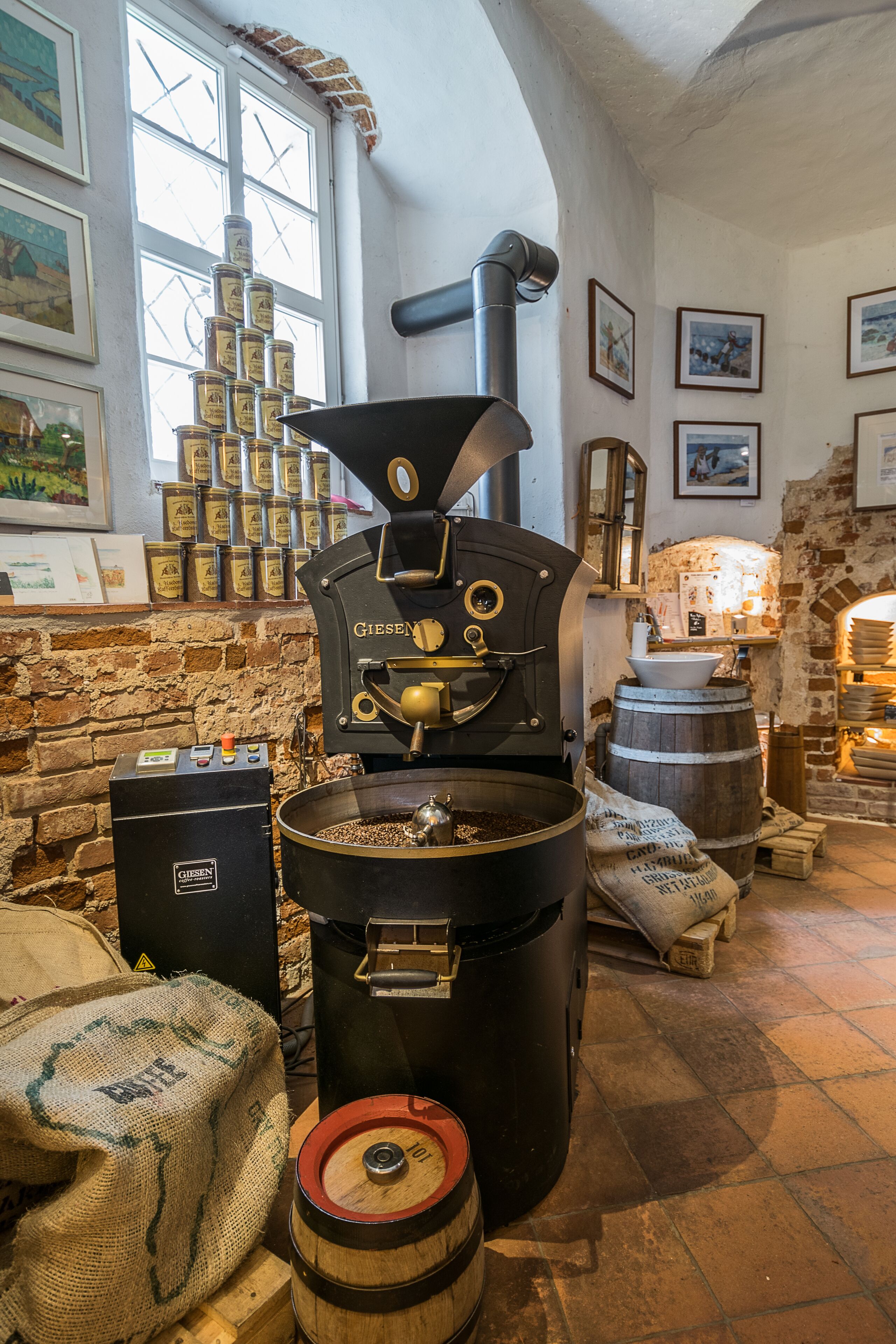A coffee roasting machine, sacks of coffee beans, and a rustic brick wall with framed pictures.