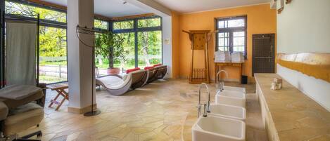 Un área de spa con suelo de piedra, varios lavabos, una zona de estar con banco y sillas, y ventanas amplias que ofrecen vistas a la vegetación.