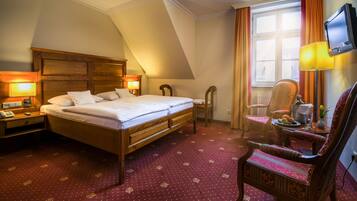 Une chambre d’hôtel avec un lit en bois, deux tables de chevet avec des lampes, une chaise, une petite table avec un vase et un téléviseur fixé au mur.