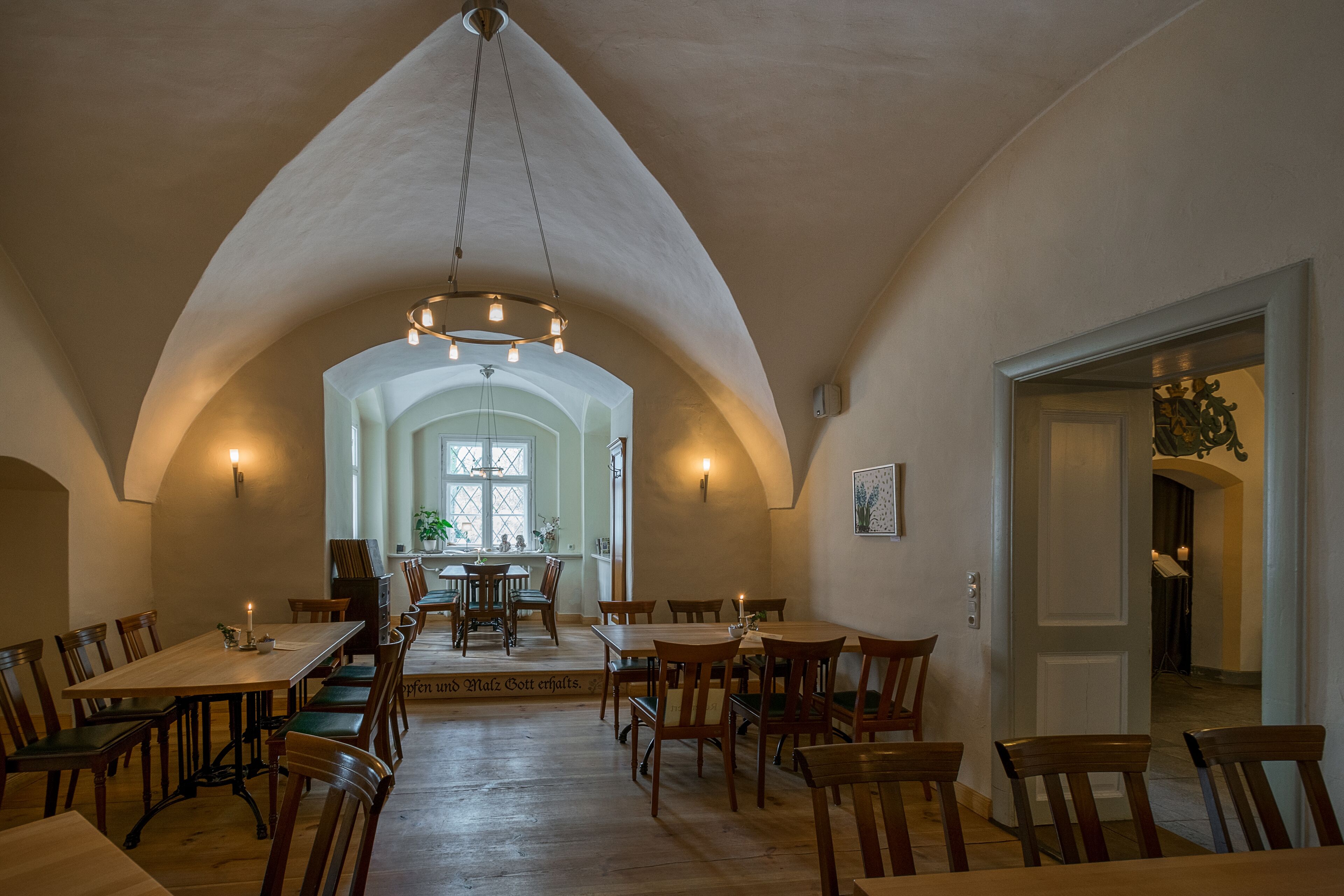 A dining area with wooden tables and chairs, a chandelier, and a high arched ceiling.