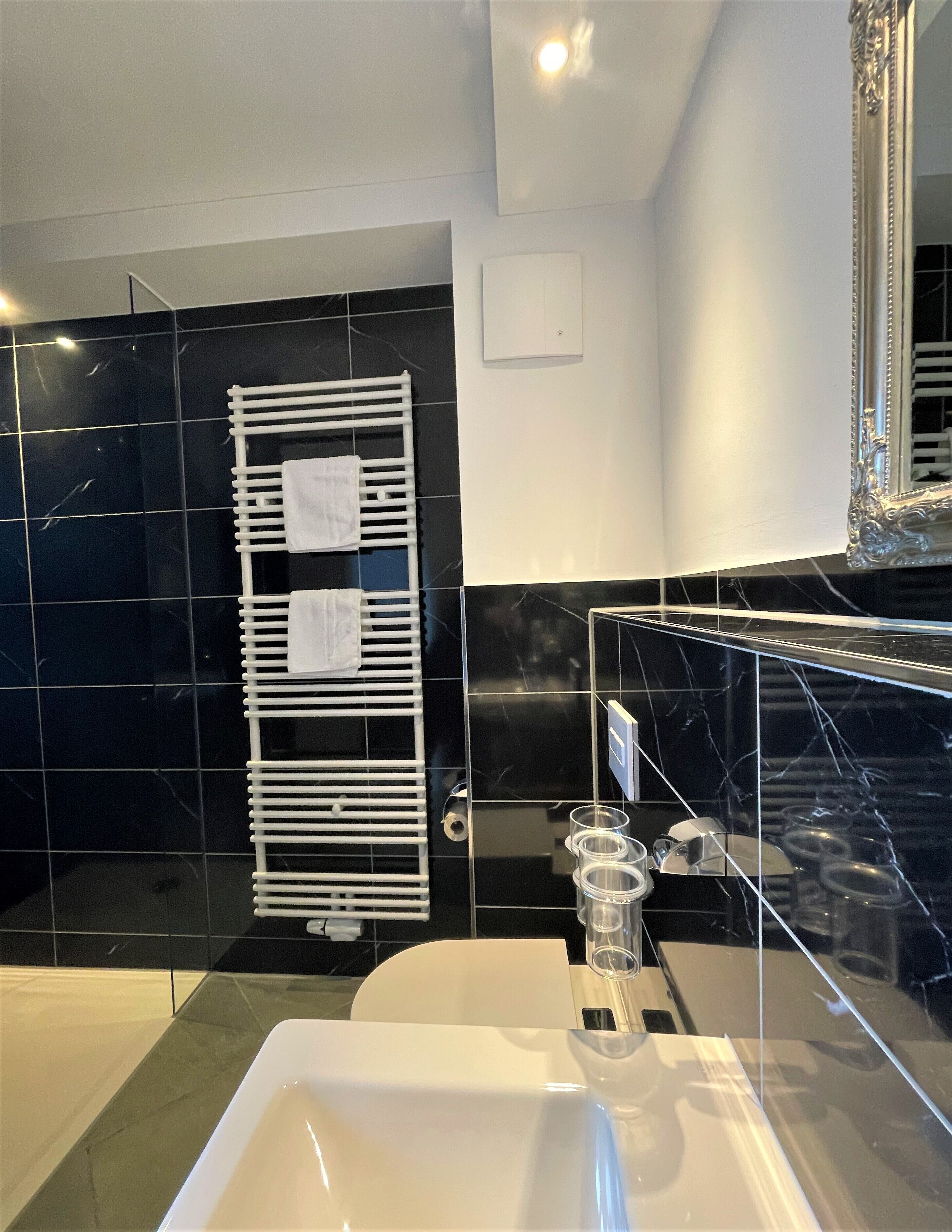 Une salle de bain moderne avec un lavabo blanc, des murs recouverts de carreaux noirs, un porte-serviettes blanc et un grand miroir.