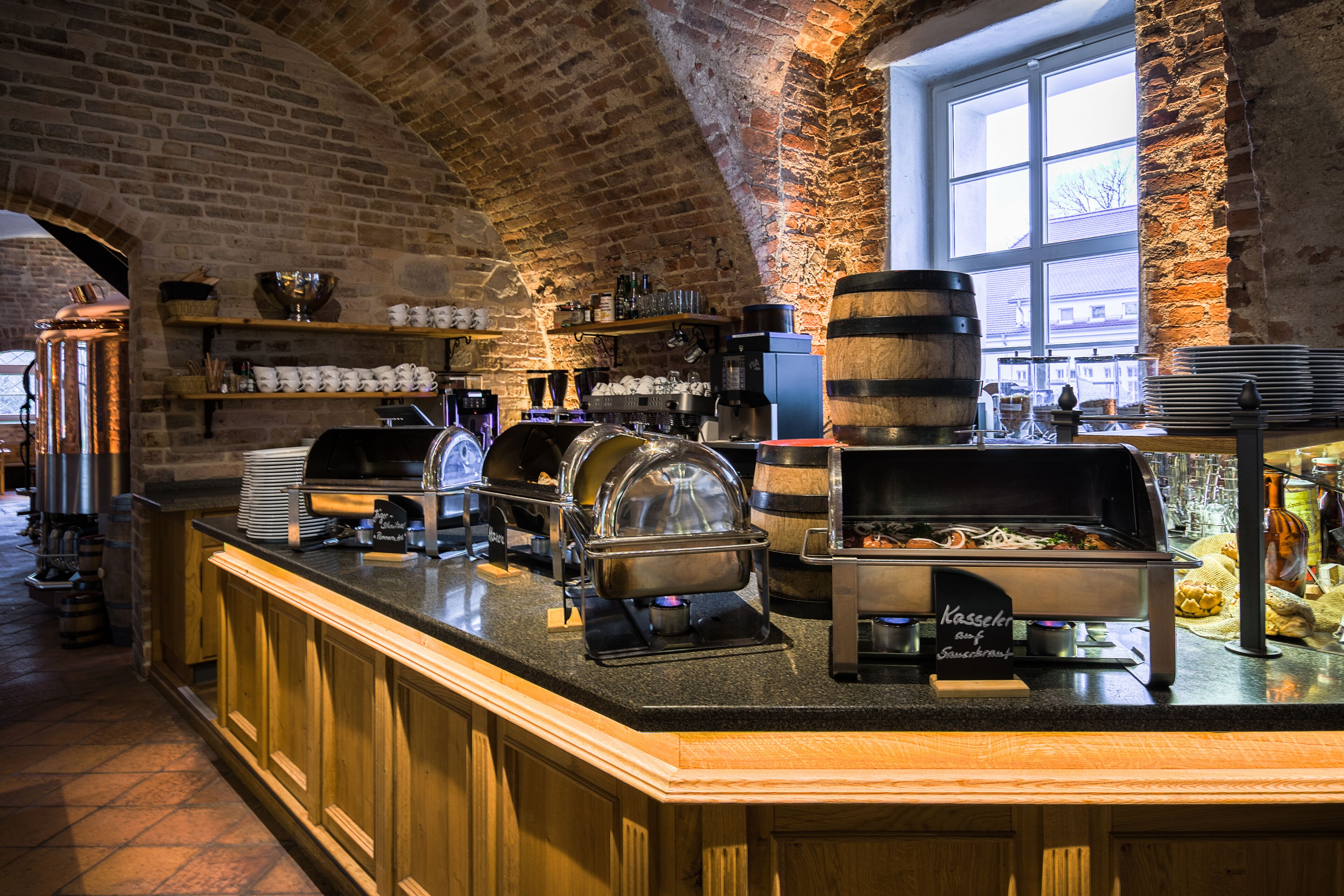 Ein reichhaltiges Buffet mit einer Kaffeestation, einer Speisenwärmeanlage und einem Getränkespender in einem raumhoch gestalteten Raum mit Backsteinwänden und einem großen Fenster.