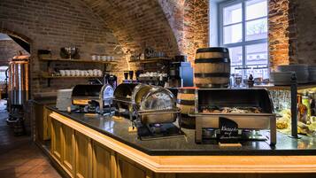 Ein reichhaltiges Buffet mit einer Kaffeestation, einer Speisenwärmeanlage und einem Getränkespender in einem raumhoch gestalteten Raum mit Backsteinwänden und einem großen Fenster.