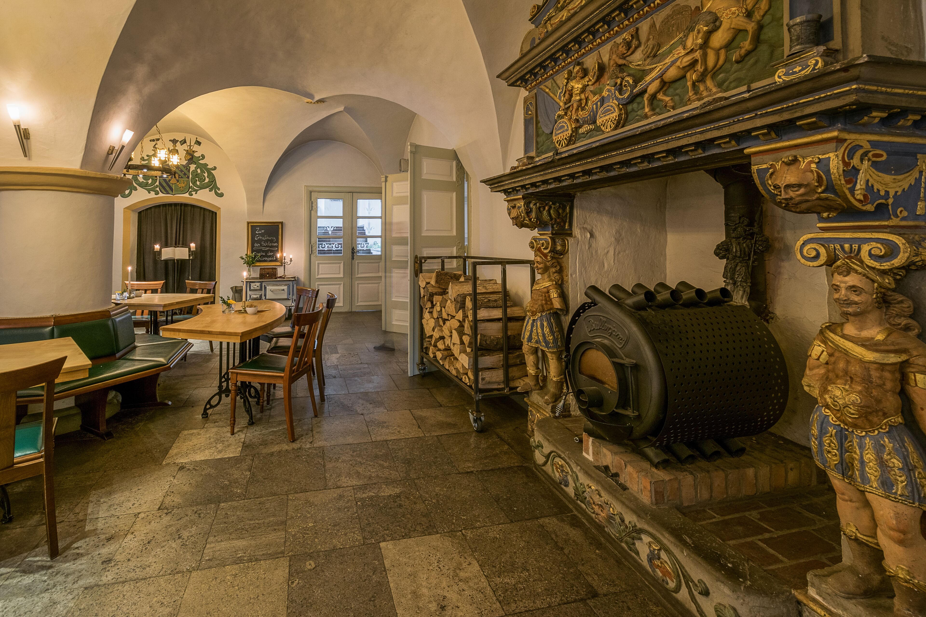 A historic interior with a stone floor, wooden furniture, a fireplace with a large barrel, and ornate carvings.