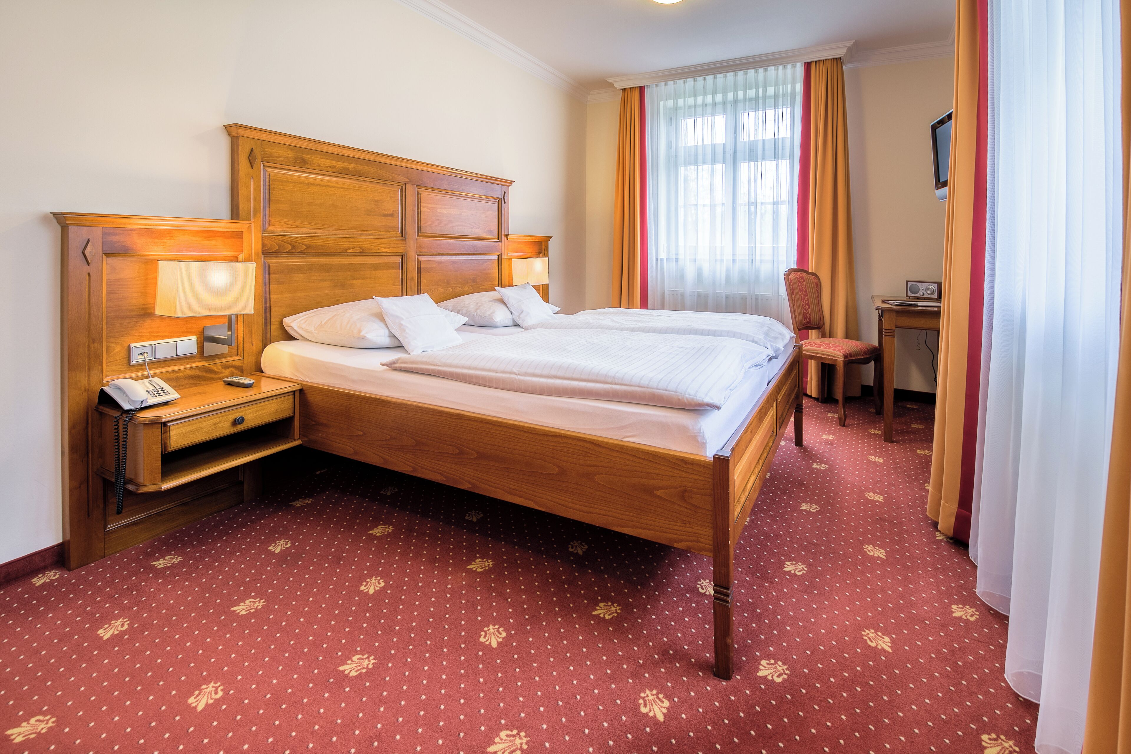 A hotel room with a large bed, a bedside table with a lamp, a chair, and a desk with a radio.