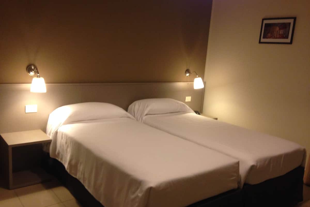 Habitación de hotel con dos camas individuales, una mesita de noche y lámparas fijadas a la pared.