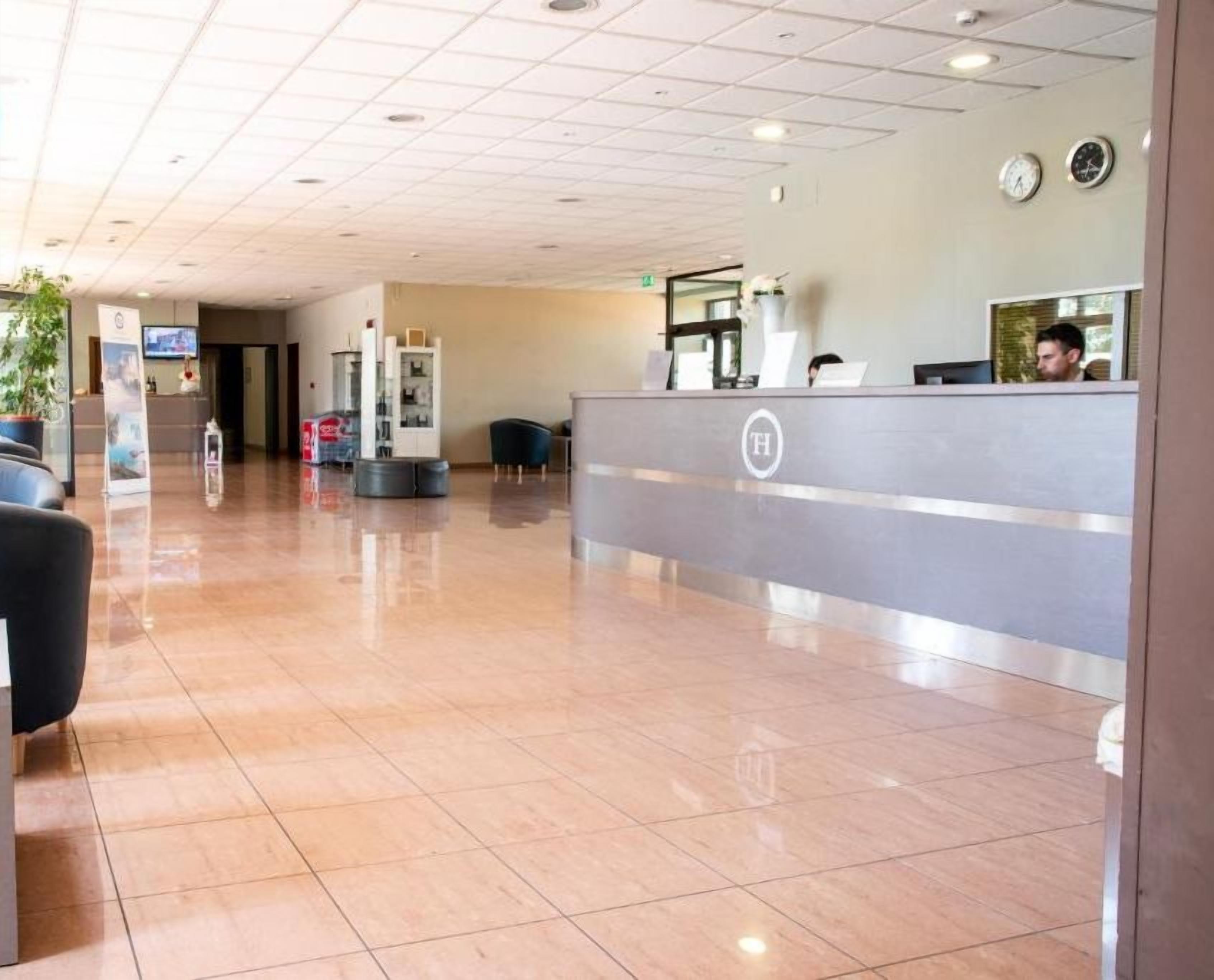 Uma área de recepção de hotel com design moderno, apresentando um balcão de atendimento, área de estar com cadeiras e piso revestido de azulejos.