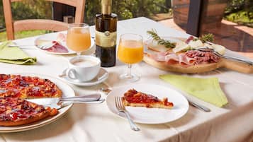 Una tavola apparecchiata per la colazione con pizza, una fetta di pizza, una bottiglia di olio d'oliva, bicchieri di succo d'arancia e un piatto di salumi.